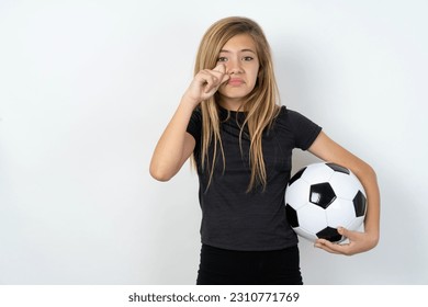 Disappointed dejected beautiful caucasian teen girl wearing sportswear holding a football ball over white wall wipes tears stands stressed with gloomy expression. Negative emotion - Powered by Shutterstock