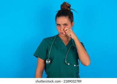 Disappointed dejected beautiful doctor woman wearing medical uniform over blue background wipes tears stands stressed with gloomy expression. Negative emotion