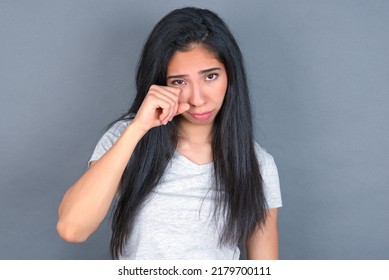 Disappointed dejected young beautiful brunette woman wearing white T-shirt over white wall wipes tears stands stressed with gloomy expression. Negative emotion