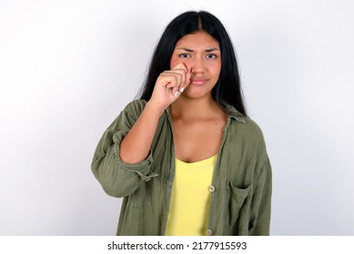 Disappointed dejected Young hispanic woman wearing green jacket over white background wipes tears stands stressed with gloomy expression. Negative emotion
