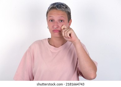 Disappointed dejected young woman with short hair wearing pink t-shirt over white background wipes tears stands stressed with gloomy expression. Negative emotion