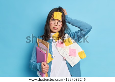 Disappointed forgetful young woman tired of cramming for exam keeps hand on head and looks stressed at camera poses with papers and stickers isolated over blue background has very tired look
