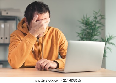 Disappointed Freelancer At Home Office Desk Covering Face With Hand In Front Of Laptop Computer. Job Problems And Professional Hardship Concept.