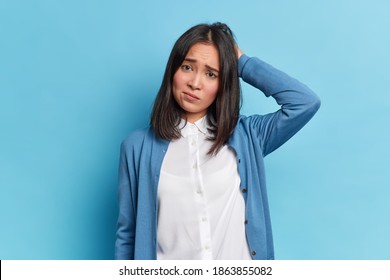 Disappointed grumpy brunette young woman with eastern appearance scratches head frowns face looks unhappily at camera wears white shirt and blue jumper poses indoor. Negative emotions concept