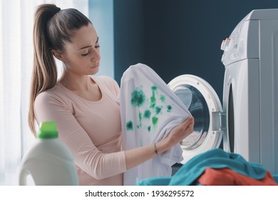Disappointed housewife standing next to the washing machine and holding stained clothes after doing laundry