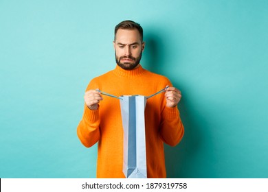 Disappointed Man Open Shopping Bag And Dislike Gift, Frowning Displeased, Standing In Orange Sweater Against Turquoise Background