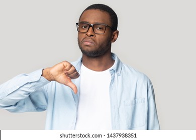 Disappointed millennial black man in glasses look at camera isolated on grey studio background show thumb down gesture, upset african American make use finger feel disappointed with service