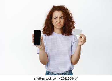 Disappointed Redhead Woman Showing Mobile Phone Screen, Credit Card, Grimacing Upset, Dislike And Disapprove Smth Bad, Standing Over White Background