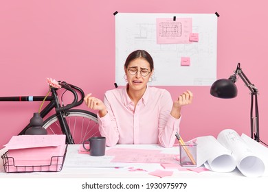 Disappointed sad female office worker feels despair cannot finish new building design project work surrounded with papers at desktop isolated on pink background sketch and bike behind. Working concept