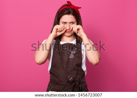 Disappointed upset housewife touching her face with hands, wiping tears out, crying, wearing brown apron, white t shirt and red headband, being in bad mood, tired of household chores. Routine concept,