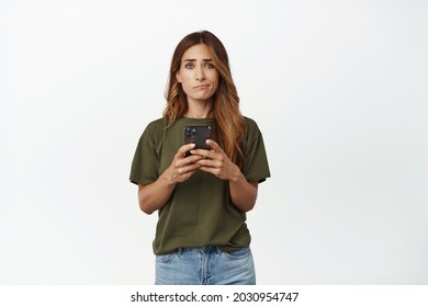 Disappointed And Upset Middle Aged Woman Holding Smartphone, Furrow Eyebrows And Smirk With Sad Face, Smth Upsetting On Mobile Screen, Standing Against White Background