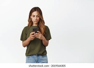 Disappointed Woman Looking At Mobile Phone With Upset Frowning Face, Not Enough Money On Bank Account On Smartphone App, Standing Displeased Against White Background