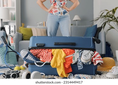 Disappointed woman packing for a journey: she is standing in front of her suitcase overfilled with clothes, travel and vacations concept