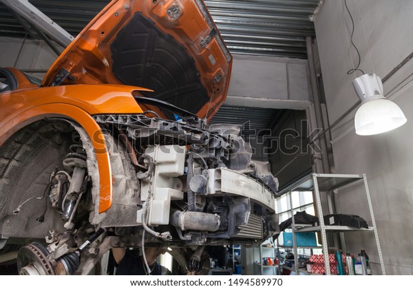 Disassembled\
orange car in the workshop raised on the lift prepared for repair:\
front end, hood and bumper removed, internal equipment, parts and\
spare parts visible. Auto service\
industry.
