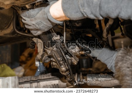Disassembly and renovation of a car's steering system in the garage. Mechanic's gloves are covered in power steering fluid as he secures the servo hoses. Exhaust pipe and shift linkage on the ground.