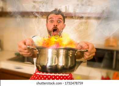 disaster home cook at kitchen- young funny and desperate man in cooking apron holding pot in flames in stress and fear making a mess of fire and smoke with food burning