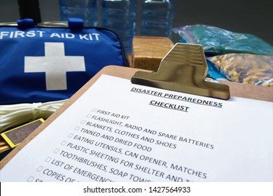 Disaster Preparedness Checklist On A Clipboard With Disaster Relief Items In The Background.Such Items Would Include A First Aid Kit,flashlight,tinned Food,water,batteries And Shelter.