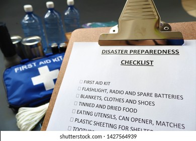 Disaster Preparedness Checklist On A Clipboard With Disaster Relief Items In The Background.Such Items Would Include A First Aid Kit,flashlight,tinned Food,water,batteries And Shelter.