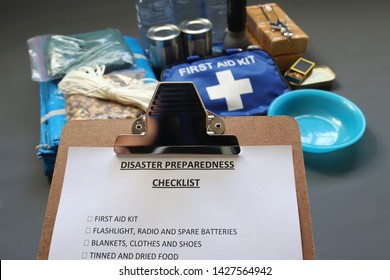 Disaster Preparedness Checklist On A Clipboard With Disaster Relief Items In The Background.Such Items Would Include A First Aid Kit,flashlight,tinned Food,water,batteries And Shelter.