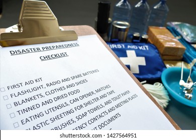 Disaster Preparedness Checklist On A Clipboard With Disaster Relief Items In The Background.Such Items Would Include A First Aid Kit,flashlight,tinned Food,water,batteries And Shelter.