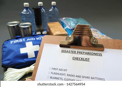 Disaster preparedness checklist on a clipboard with disaster relief items in the background.Such items would include a first aid kit,flashlight,tinned food,water,batteries and shelter.Disaster plan.