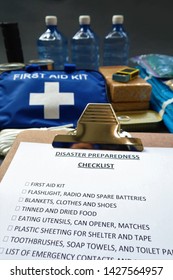 Disaster Preparedness Checklist On A Clipboard With Disaster Relief Items In The Background.Such Items Would Include A First Aid Kit,flashlight,tinned Food,water,batteries And Shelter.