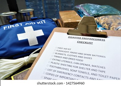 Disaster Preparedness Checklist On A Clipboard With Disaster Relief Items In The Background.Such Items Would Include A First Aid Kit,flashlight,tinned Food,water,batteries And Shelter.