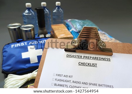 Disaster preparedness checklist on a clipboard with disaster relief items in the background.Such items would include a first aid kit,flashlight,tinned food,water,batteries and shelter.Disaster plan.