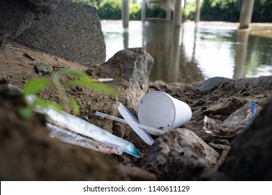 Discarded Food Containers Litter The Area Under A Bridge Near A River With Trash That Includes Plastic Straws, Plastic Lids, A Styrofoam Cup And Other Plastic Food Waste, Contributing To Pollution.