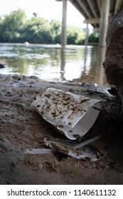 Discarded Food Containers Litter The Area Under A Bridge Near A River With Trash That Includes Plastic Straws, Plastic Lids, A Styrofoam Cup And Other Plastic Food Waste, Contributing To Pollution.