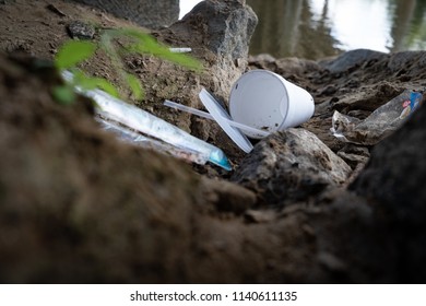 Discarded Food Containers Litter The Area Under A Bridge Near A River With Trash That Includes Plastic Straws, Plastic Lids, A Styrofoam Cup And Other Plastic Food Waste, Contributing To Pollution.