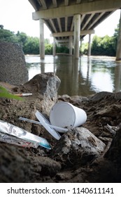 Discarded Food Containers Litter The Area Under A Bridge Near A River With Trash That Includes Plastic Straws, Plastic Lids, A Styrofoam Cup And Other Plastic Food Waste, Contributing To Pollution.