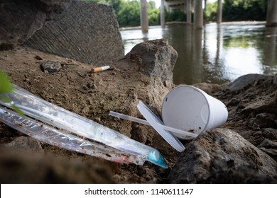 Discarded Food Containers Litter The Area Under A Bridge Near A River With Trash That Includes Plastic Straws, Plastic Lids, A Styrofoam Cup And Other Plastic Food Waste, Contributing To Pollution.