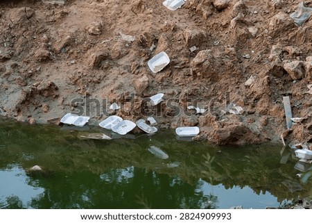 Discarded plastic and Styrofoam containers float in polluted water beside a muddy canal bank in Hanoi, Vietnam, highlighting urban waste and environmental neglect.