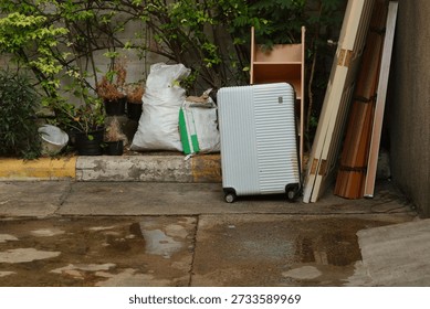 A discarded white hardshell rolling suitcase with vertical ridges stands on a wet concrete surface. The suitcase shows signs of wear and dirt, particularly yellowish-brown stains along its bottom edge and sides. Behind the suitcase a wooden shelf stands empty, to its right, wooden doors and furniture components are leaning against a textured wall. To its immediate left are two large, stuffed sacks along with several plants, some thriving with green leaves and others dry and brown, all set against a backdrop of a cinder block wall.