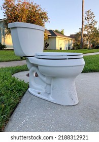 Discarded White Toilet Standing Curbside In Front Of House In Early Morning Sun. Suburban Residential Neighborhood Street Scene.