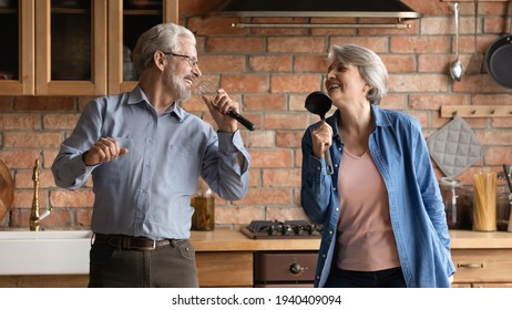 Disco 60s. Overjoyed older senior husband wife having fun sing song at kitchen hold imaginary microphones of tools utensils. Energetic creative aged spouses listen to music enjoy karaoke funny cooking