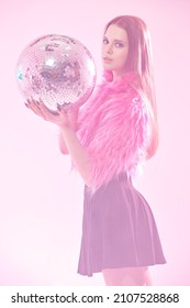 Disco, Party Girl. An Attractive Sexy Girl In School Clothes, A Glamorous Fuchsia Fur Jacket And With A Sparkling Disco Ball In Her Hands Posing In Pink Light. 