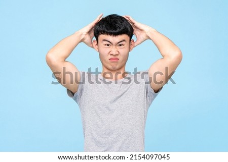 Discontent Asian man touching head with furious expression while standing against light blue isolated background in studio
