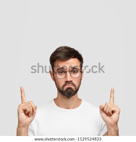 Discontent bearded male costumer can`t choose suitable outfit in shop, purses lips with displeasure, points upwards with both index fingers, has gloomy expression, isolated on white studio wall