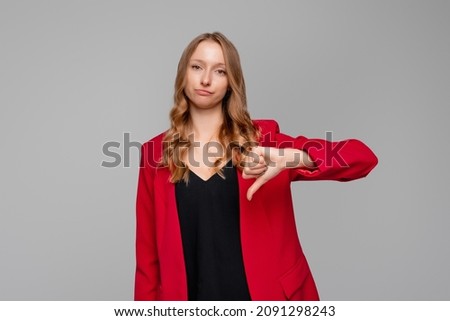 Discontent blonde woman shows disapproval sign, keeps thumb down, expresses dislike, frowns face in discontent, standing in red blazer against gray background. Body language concept