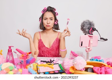 Discontent brunette Asian woman looks with indignant expression holds cosmetic brush for applying beauty powder makes hairstyle applies hair rollers records beauty blog on smartphone webcam. - Powered by Shutterstock