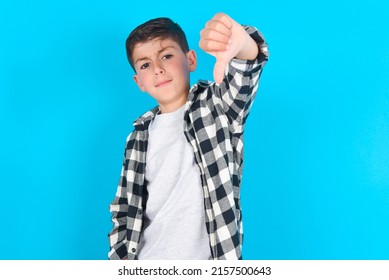 Discontent Caucasian Kid Boy Wearing Plaid Shirt Over Blue Background Shows Disapproval Sign, Keeps Thumb Down, Expresses Dislike, Frowns Face In Discontent. Negative Feelings.