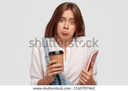 Discontent lovely European student purses lips, looks with negative facial expression, dressed in fashionable clothes, holds book and takeaway coffee, has clueless look at camera, poses at studio