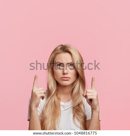 Discontent pretty blonde woman points with fore fingers above head at blank copy space, looks in bewilderment upwards as demonstrates something strange, isolated over pink studio background.