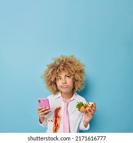 Discontent Thoughtful Curly Woman Uses Cellphone Eats Tasty Hot Dog Wears White Formal Shirt Smeared With Ketchup Prefers Fast Food For Quick Snack Feels Unhappy Because Of Bad Eating Habits