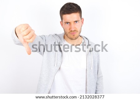 Discontent young caucasian man wearing casual clothes over white background shows disapproval sign, keeps thumb down, expresses dislike, frowns face in discontent. Negative feelings.