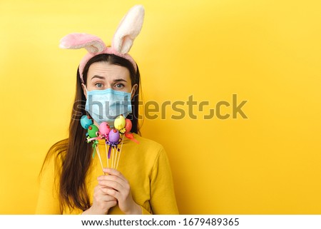 Discontent young woman with virus mask and looks unhappily, has spoiled day, poses with small Easter eggs, wears spectacles, rabbit fluffy ears, poses. Concept holiday, coronavirus, virus protection