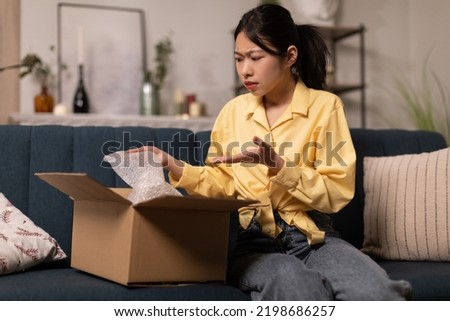 Discontented Asian Lady Buyer Unpacking Cardboard Box With Bad Product Sitting On Sofa At Home. Negative Feedback, Delivery Service And Shopping Problem Concept