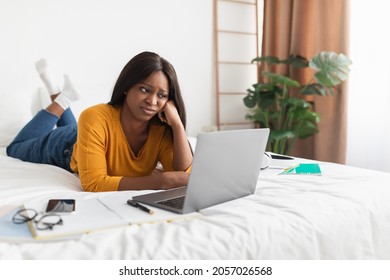 Discontented Black Millennial Woman Looking At Laptop Computer Lying In Bed, Being Bored And Unhappy While Working Or Learning Online, Browsing Internet At Home. People And Gadgets Concept